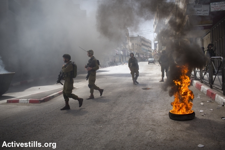 Israeli soldiers walk near a burning tire during clashes in Hebron. (photo: Activestills)