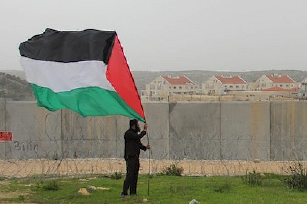 A Palestinian stakes a giant flag in front of the Separation Wall in the village of Bi'lin with a Jewish settlement in the background. (photo: Omar Rahman)