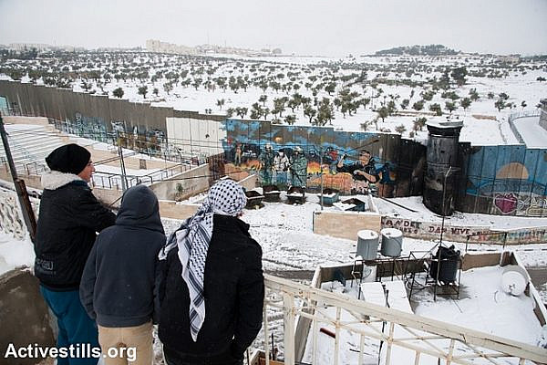 Palestinian youth stand on a rooftop in Aida Refugee Camp overlooking the Israeli separation wall and snow-covered olive groves on the other side, Bethlehem, West Bank, December 14, 2013. (photo: Activestills)