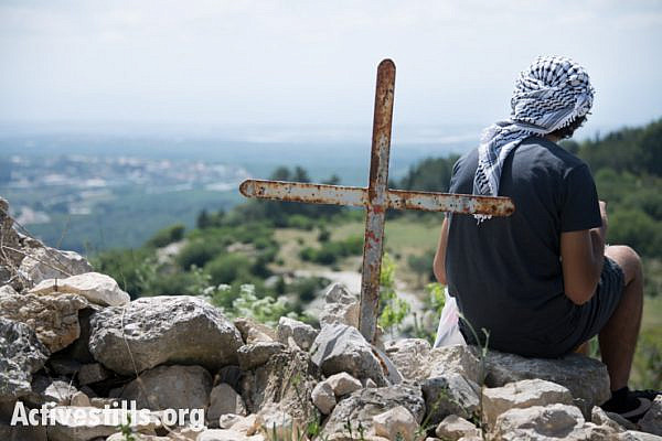 A youth sits near a cross overlooking the surrounding countryside in the displaced Palestinian village of Iqrit in northern Israel, April 21, 2014. Iqrit's original inhabitants were forcibly evacuated in the Nakba of 1948. Though the Israeli high court granted the residents, who are Palestinian citizens of Israel, the right to return to their homes in 1951, the military destroyed the village and has since prevented their return. Only the village's church and cemetery remained intact, and are still used by village residents while they campaign for a full return.