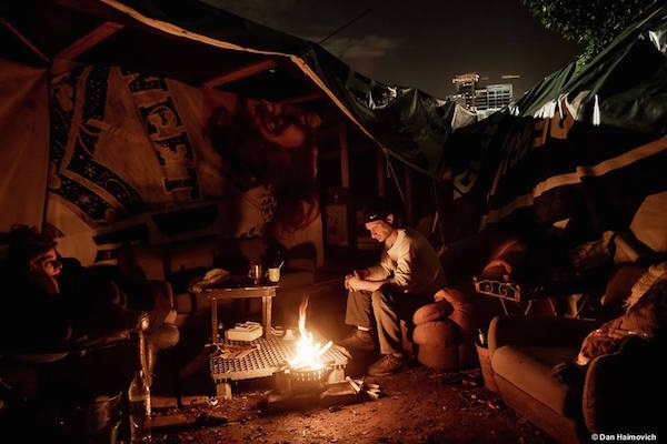 A man sits around a small fire at the Arlozorov encampment in Tel Aviv. (Photo by Dan Haimovich)