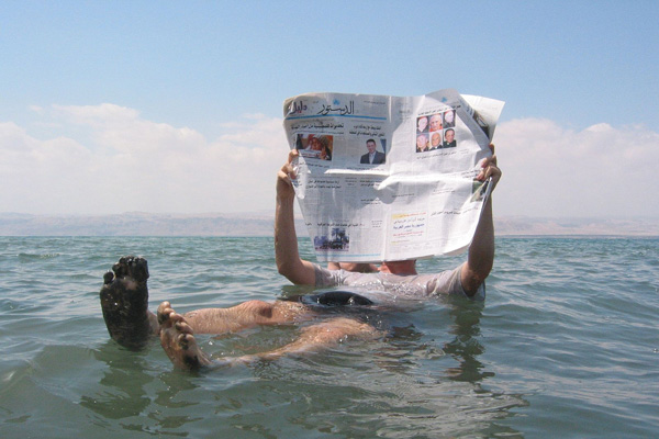 A man reads a newspaper while floating in the Dead Sea. (Author unknown, WikiCommons)