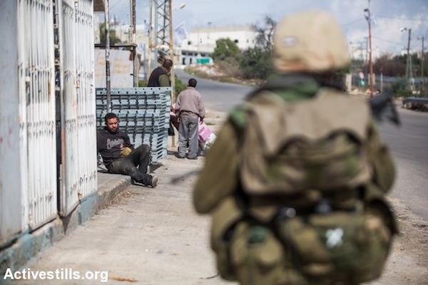 A Palestinian laborer rests on the side of a street in occupied Hebron as an Israeli soldier patrols, October 29, 2015. (Yotam Ronen/Activestills.org)