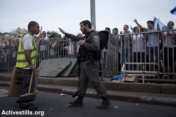 An Israeli police officer argues with an Arab sanitation worker in Jerusalem as right-wing Jewish Israelis stand behind a barricade during the nationalist and often violently racist “March of the Flags” parade, Jerusalem, May 28, 2014. (Oren Ziv/Activestills.org)