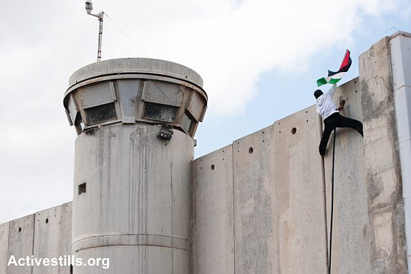 A Palestinian scales the Israeli separation wall and plants his flag during a protest at Bethlehem checkpoint, March 30, 2012. (Activestills.org)