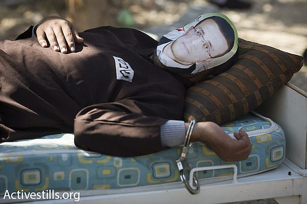 A Palestinian man wearing the likeness of hunger striking Palestinian journalist Muhammad al-Qiq lies shackled to a bed. Al-Qiq has been on hunger strike for 83 days in protest of his administrative detention. Bil’in, February 19, 2016. (Oren Ziv/Activestills.org)
