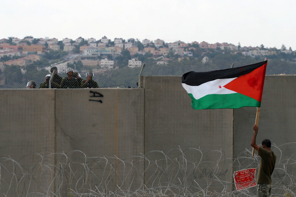 A Palestinian protester holds a flag as Israeli soldiers look on from beyond the separation wall, Bil’in. (Issam Rimawi/Flash90)