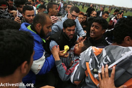 Palestinian men tend to a wounded protester during the Great Return March in Gaza, east of Jabaliya. March 30, 2018. (Mohammed Zaanoun / Activestills.org)
