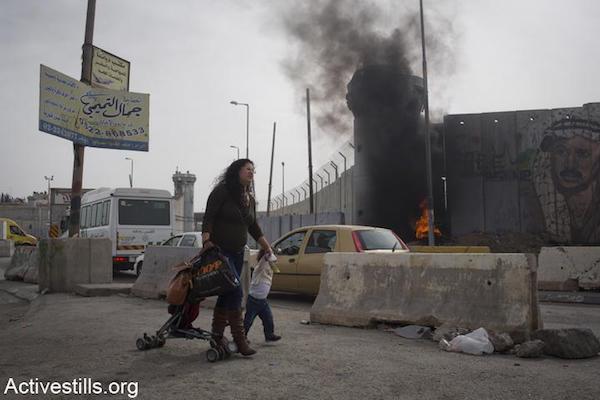 A Palestinian mother and her child walk past the Israeli army's Qalandiya checkpoint as clashes take place. (File photo by Oren Ziv/Activestills.org)