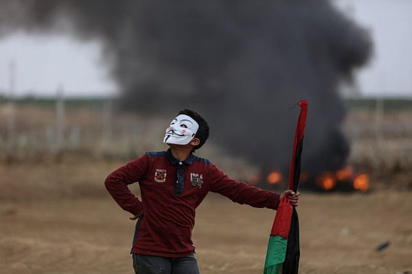 A young Palestinian protester wearing a Guy Fawkes mask holds a Palestinian flag and looks on during clashes with Israeli soldiers in eastern Gaza City, May 4, 2018. (Wissam Nassar/Flash90)