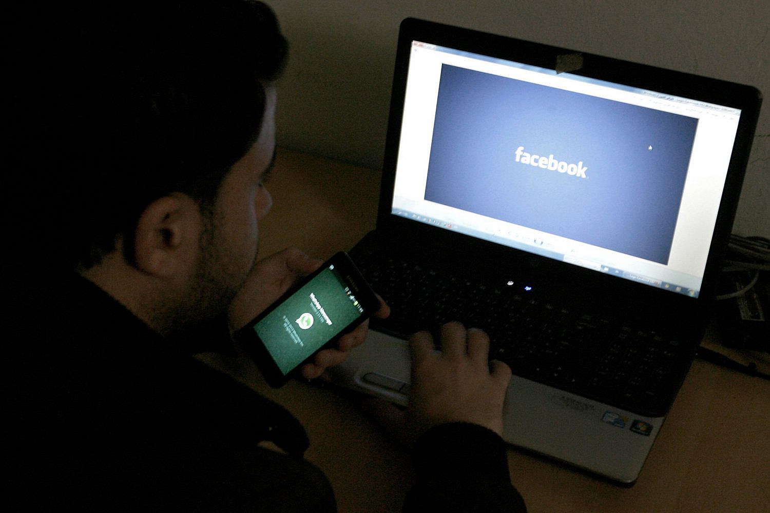 A Palestinian youth holds a phone displaying WhatsApp in front of a computer with Facebook in Rafah in the southern Gaza Strip on February 26, 2014. (Abed Rahim Khatib/Flash90)