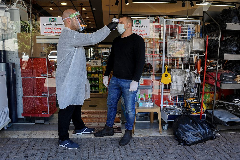 A man checks the temperature of a customer to identify if he has a fever, in the northern Arab town of Deir al-Asad, April 18, 2020. (Basel Awidat/Flash90)