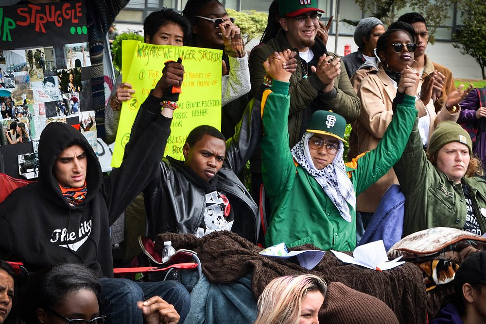 (Center left to right) Sachiel Rosen, Hassani Bell, Ahkeel Mestayer, and Julia Retzlaff, four students referred to as the Third World Liberation Front 2016, who were on a hunger strike to defend the funding of the San Francisco State College of Ethnic Studies, raise their joined hands during an emergency press conference in the Quad, May 9, 2016. (Melissa Minton/CC BY 2.0)
