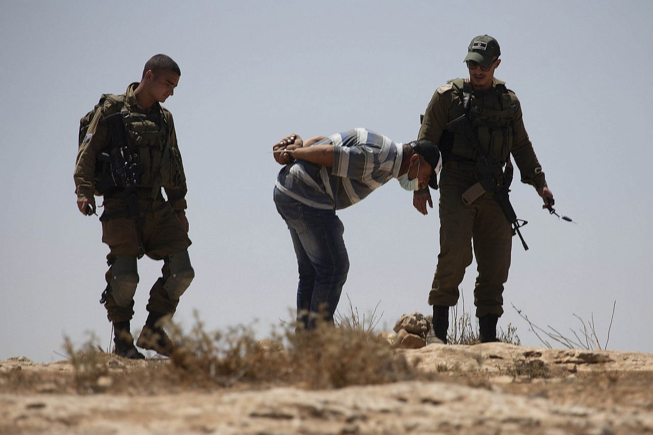 An arrested Palestinian stands handcuffed next to Israeli soldiers during a demonstration near a-Tuwani in the South Hebron Hills against new structures built in the nearby Israeli settlement of Avigail, August 20, 2021. (Oren Ziv/Activestills)