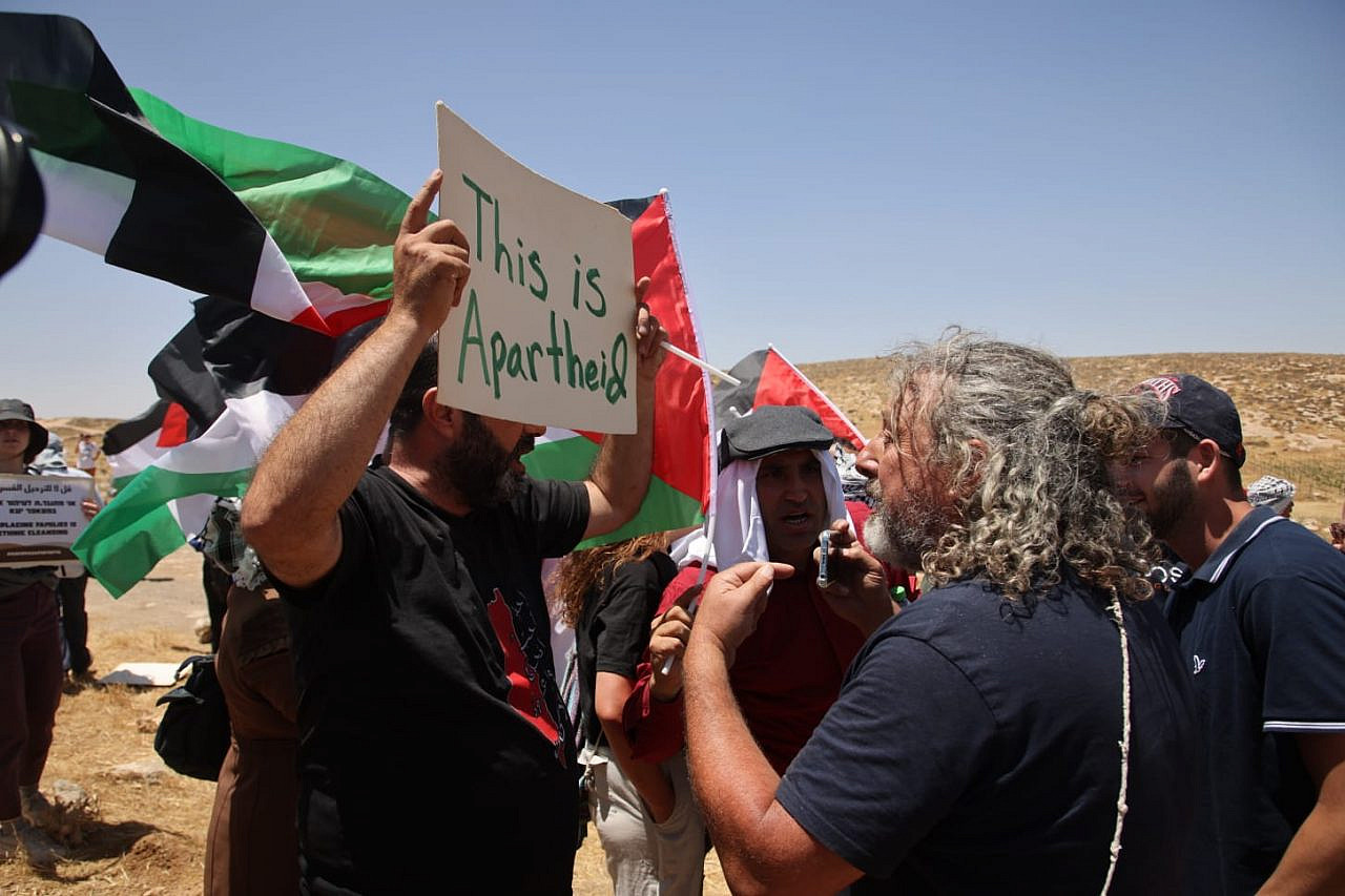 A Palestinian protestor holds a sign that says "This is Apartheid" in front of Israeli soldiers and settlers at a demonstration against the expulsion of the Palestinian communities who live inside what Israel calls Firing Zone 918, Masafer Yatta, occupied West Bank, June 10, 2022. (Oren Ziv)