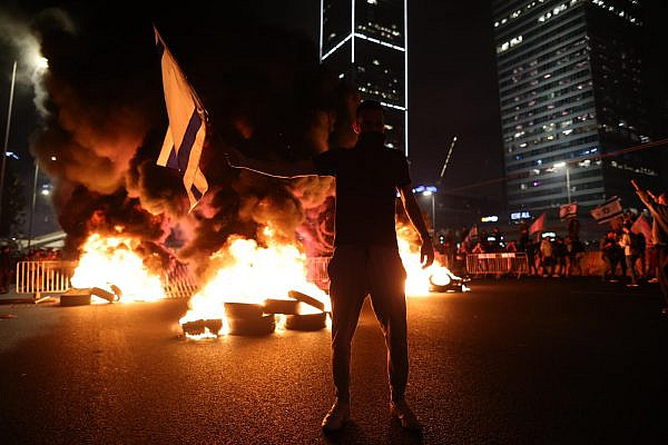 A protester stands in front of burning tires on the Ayalon highway, Tel Aviv. Tens of thousands of demonstrators took to the streets following Prime Minister Benjamin Netanyahu's dismissal of Defense Minister Yoav Gallant, after he called to pause the government's planned judicial overhaul, March 26, 2023. (Oren Ziv)