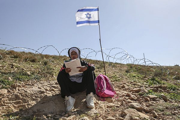 A Palestinian child reads her schoolbook in front of newly erected barbed wire fence bearing Israeli flags, which Israeli settlers put up to block the passage of Palestinian teachers and students from their homes in the village of Umm Al-Khair to their school, Masafer Yatta, occupied West Bank, April 13, 2026. (Mosab Shawer/Activestills)