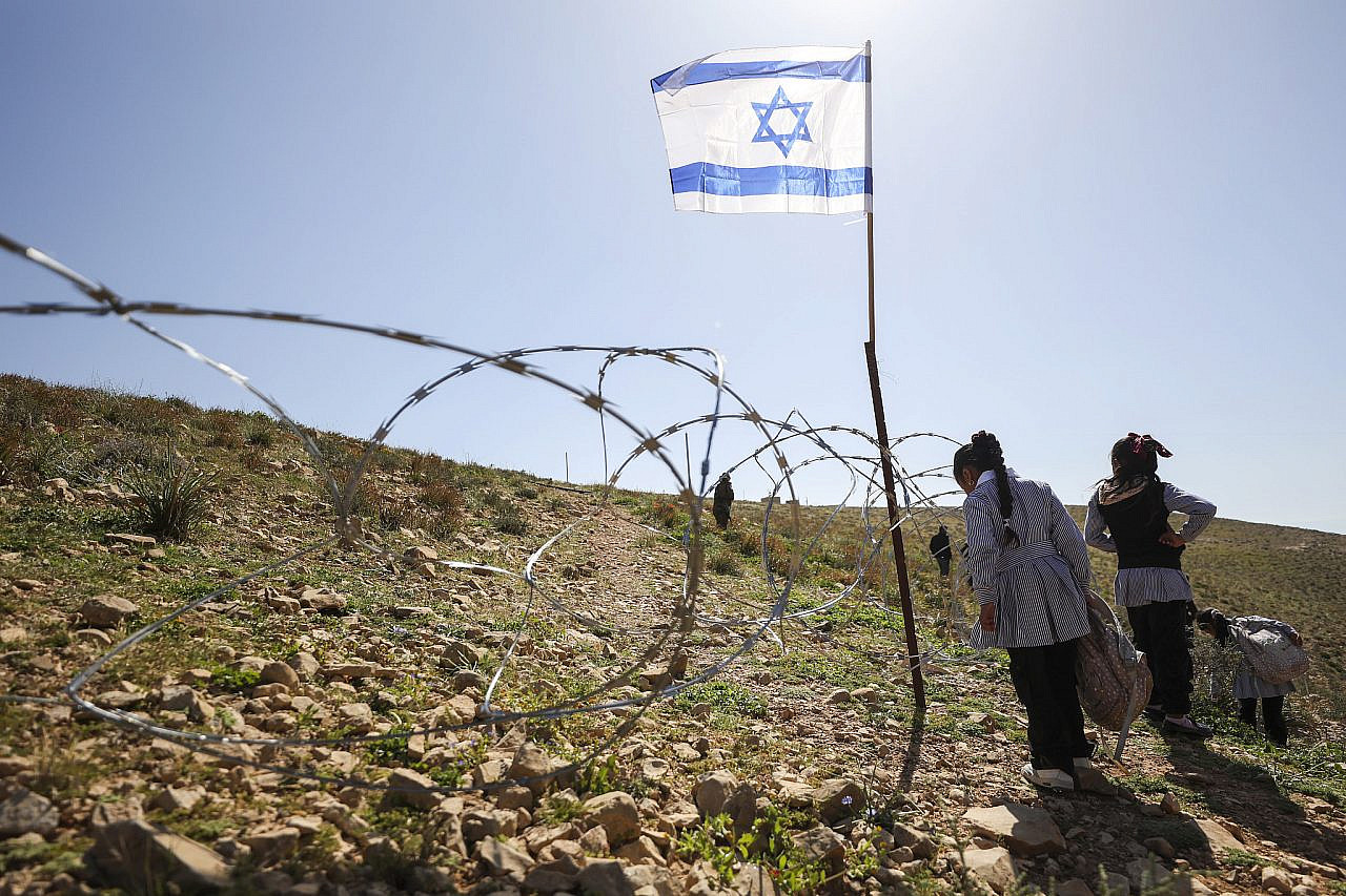 A barbed wire fence bearing Israeli flags blocks the passage of Palestinian teachers and students from the village of Umm Al-Khair to their school, Masafer Yatta, occupied West Bank, 13 April 2026. (Mosab Shawer/Activestills)