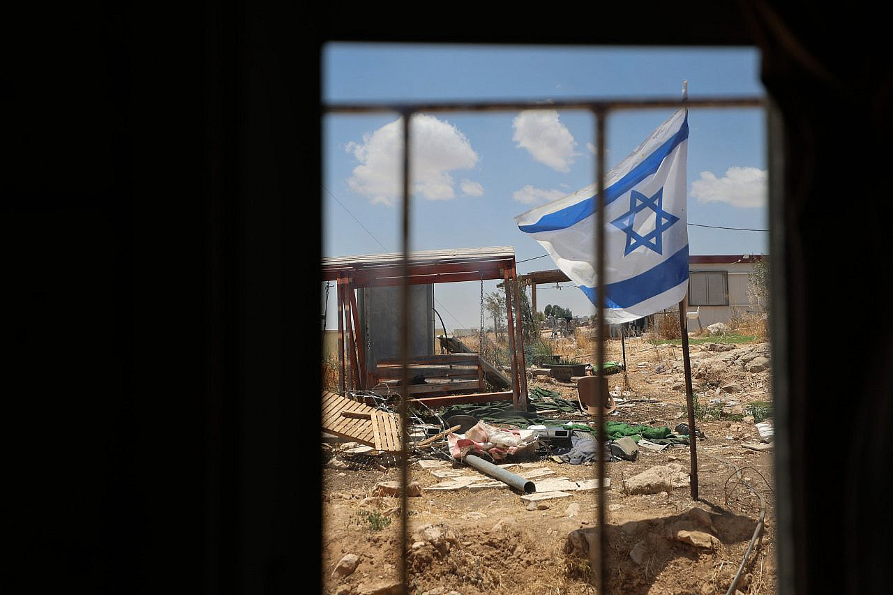 An Israeli flag erected by settlers is seen though the windows of the home of Ikhlas Al-Hathaleen. Military closure orders restricted the family from using the toilet and from feeding and milking their sheep, Umm Al-Khair, Masafer Yatta, occupied West Bank, July 6, 2026. (Activestills/Mosab Shawer)
