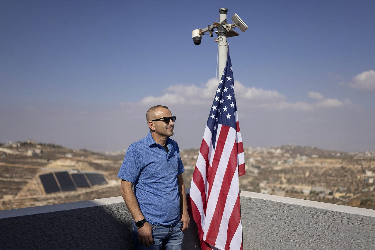 Loui Ridi stands beside an American flag on the balcony of his besieged home in the West Bank village of Qusra, August 18, 2026. (Oren Ziv)