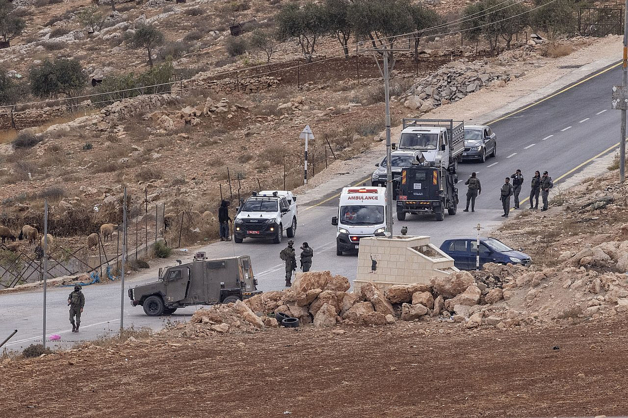 An ambulance waits at a checkpoint while trying to evacuate a Palestinian who was attacked by Israeli settlers, at the entrance to the village of Al-Mughayyir, as an Israeli settler grazes his sheep on Palestinian land to the left, occupied West Bank, October 2, 2026. (Oren Ziv)
